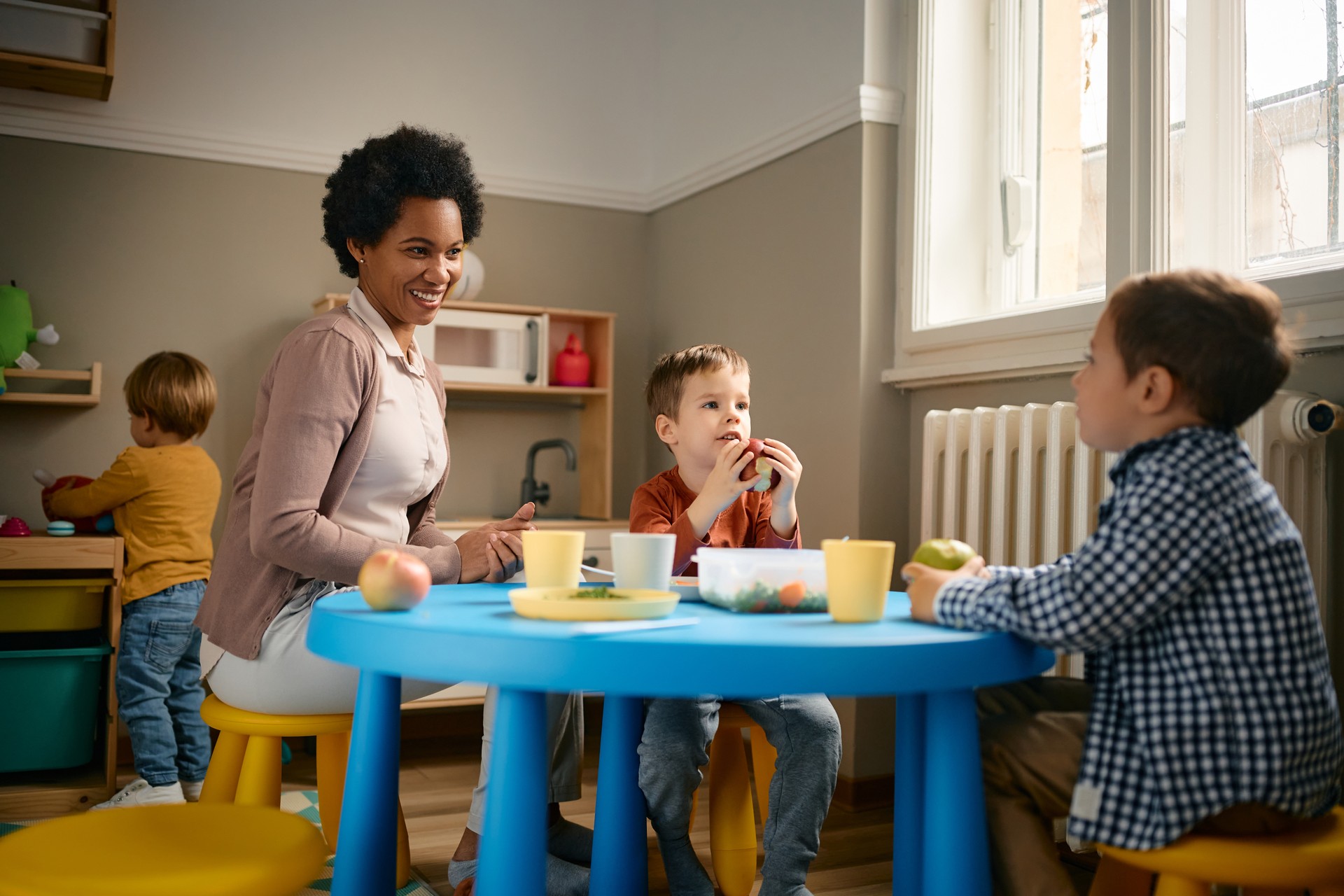 Happy black teacher and small boys enjoying during lunch break at kindergarten.